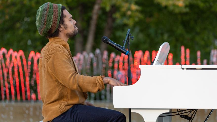 Man playing a white piano outdoors with colorful fountains in the background.