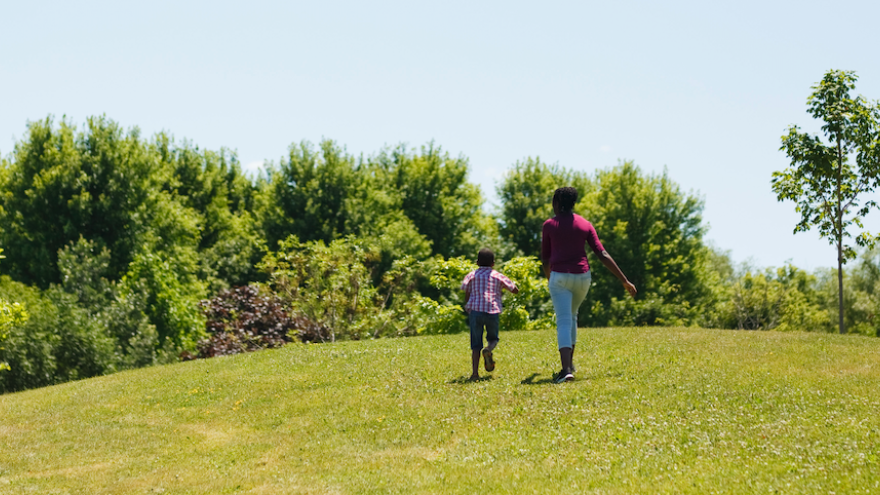 a mother and her child running on open grass towards a forest on a sunny day