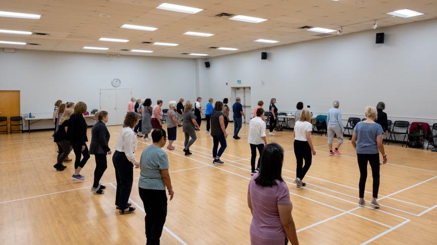 A group of adults participate in a line dance class in a large community hall 