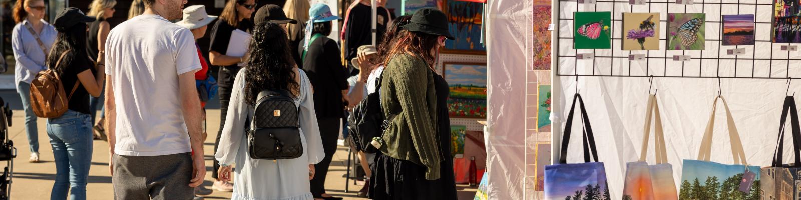 Outdoor market with people browsing art and handmade items.