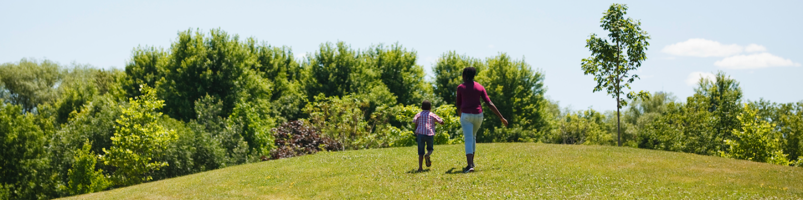 a mother and her child running on open grass towards a forest on a sunny day