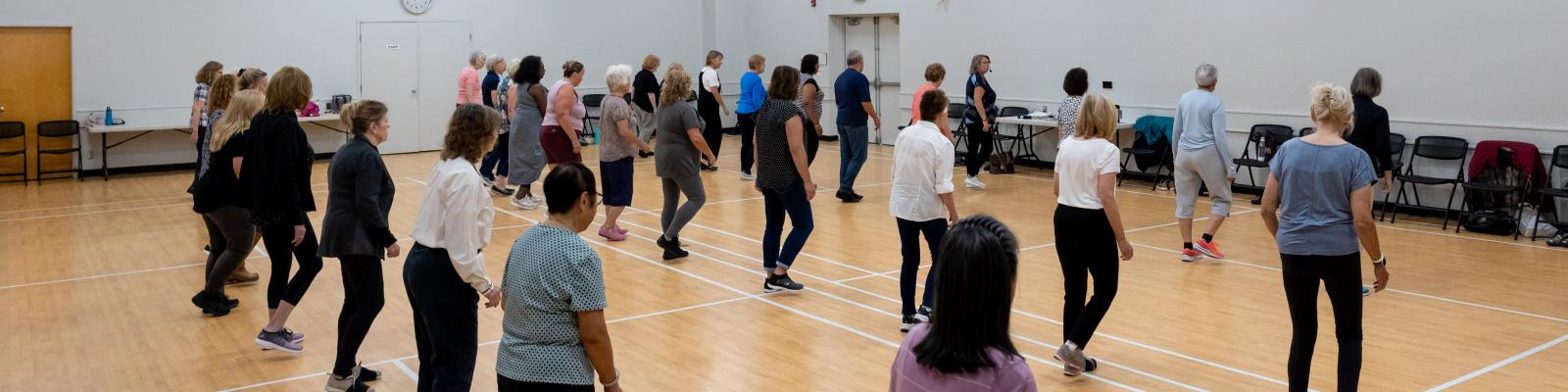 A group of adults participate in a line dance class in a large community hall 