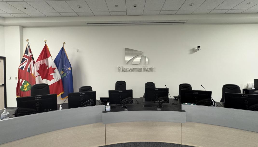 Town of Newmarket council chambers with a curved council desk, computer monitors and the Ontario, Canadian and Newmarket flags displayed beside the Newmarket logo.