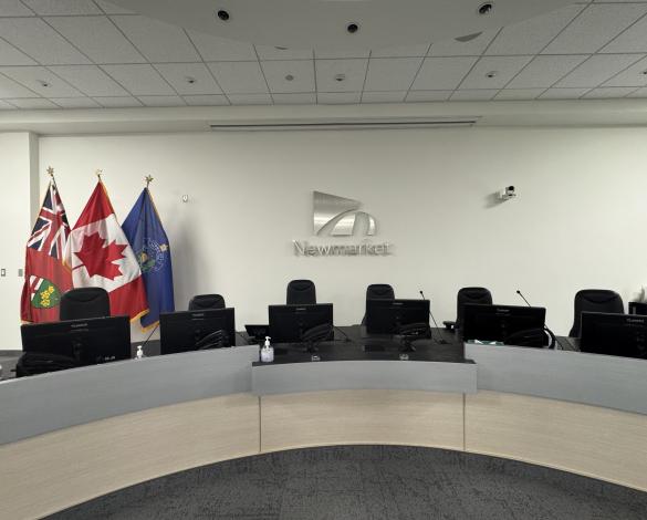Town of Newmarket council chambers with a curved council desk, computer monitors and the Ontario, Canadian and Newmarket flags displayed beside the Newmarket logo.