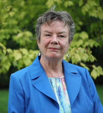 Elderly woman in a blue blazer, outdoors with green foliage.