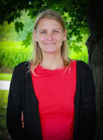 Smiling person in red shirt, standing by a tree, with greenery in the background.