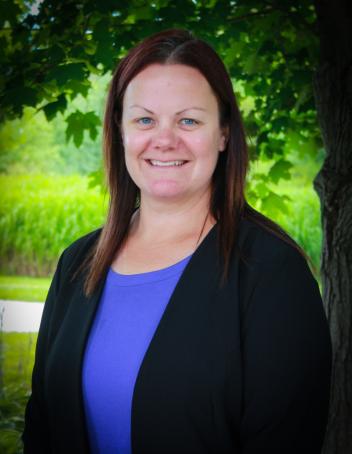 Smiling woman in a black blazer and blue shirt, standing outdoors by a tree.