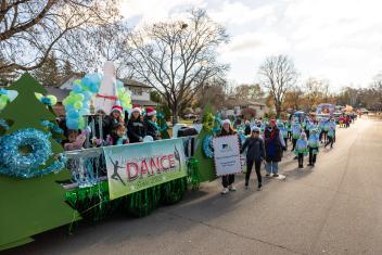 Parade with colorful floats and people walking on a tree-lined street.