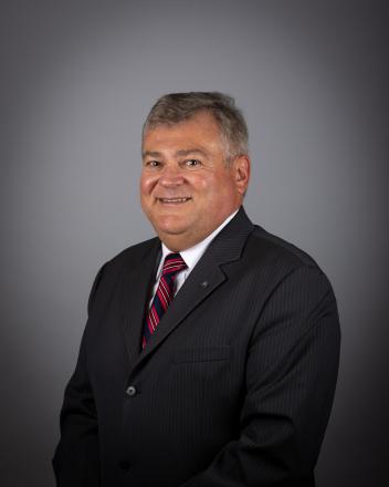 Bob Kwapis in a dark suit with a red striped tie against a gray background, looking at the camera.
