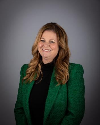 Kelly Broome smiling in a green jacket against a gray background.