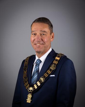 Major John Taylor with ceremonial chain necklace, smiling against a gray background.
