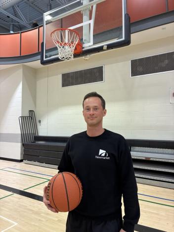 A person holds a basketball in an indoor gym, with a hoop behind them.