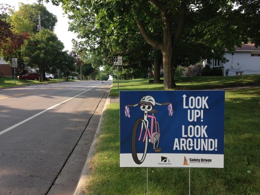Sidewalk sign with bicycle graphic under shady trees.