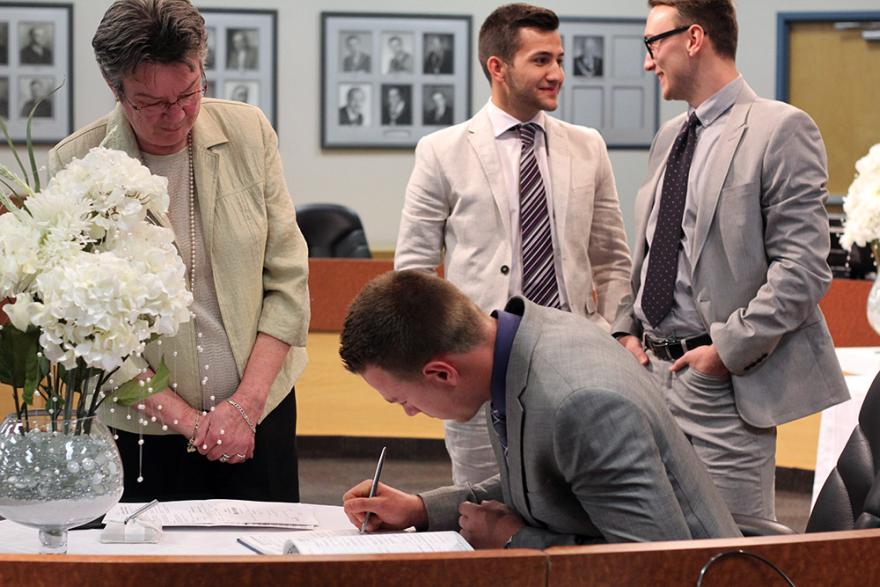 Man in suit signing document, others chatting nearby in an office setting.