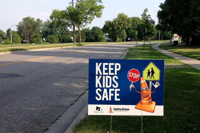 Roadside safety sign with "Keep Kids Safe" message and cartoon orange cone.