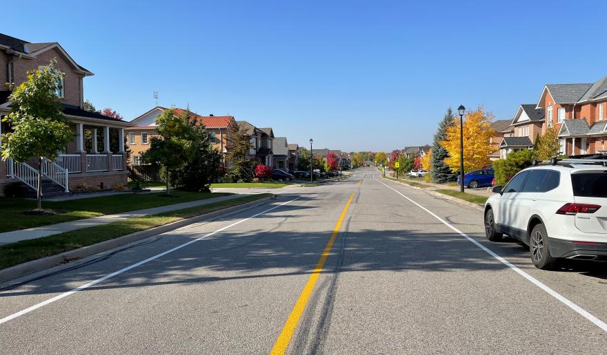 Quiet suburban street with parked cars and autumn trees under a clear blue sky.