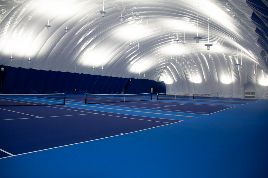 Indoor tennis courts with a domed ceiling, lit with bright lights.