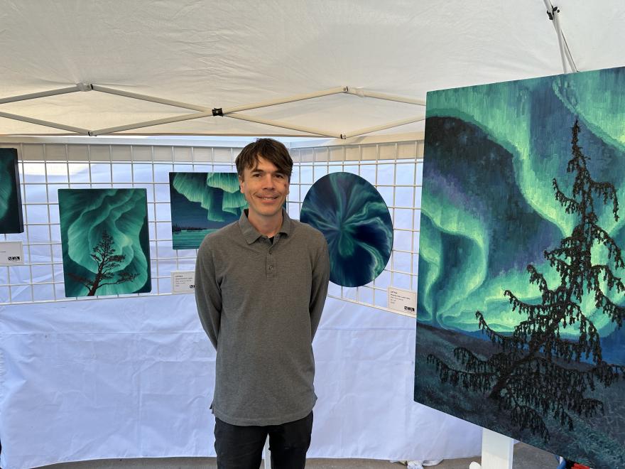 Art display under a tent with northern lights paintings; a person stands smiling.