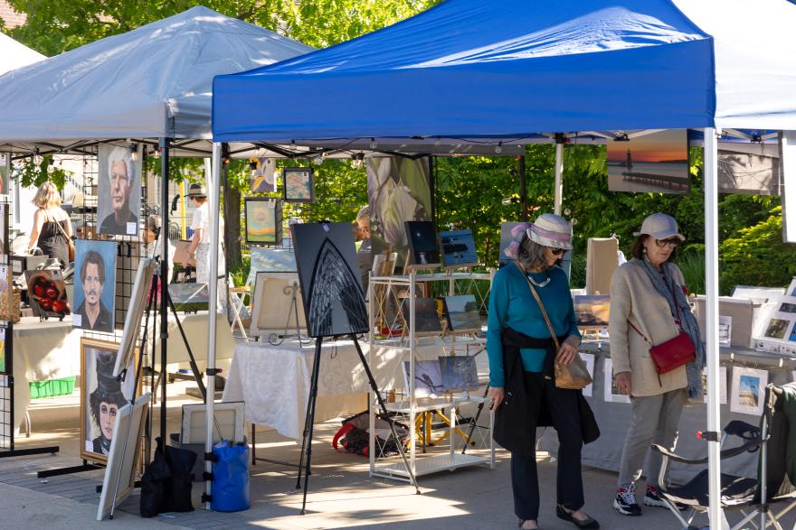 Outdoor art market with two women browsing paintings under tents.
