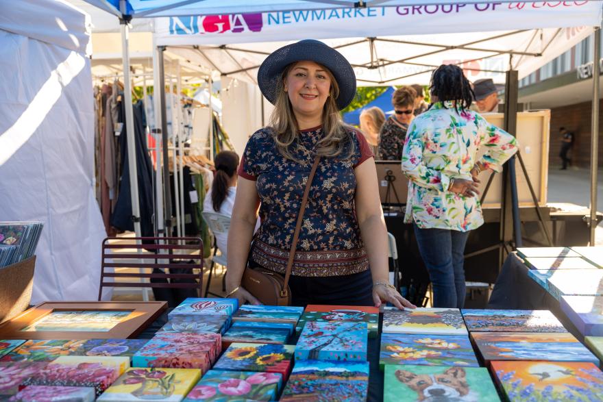 Art vendor smiling at outdoor market stall with colorful paintings displayed.