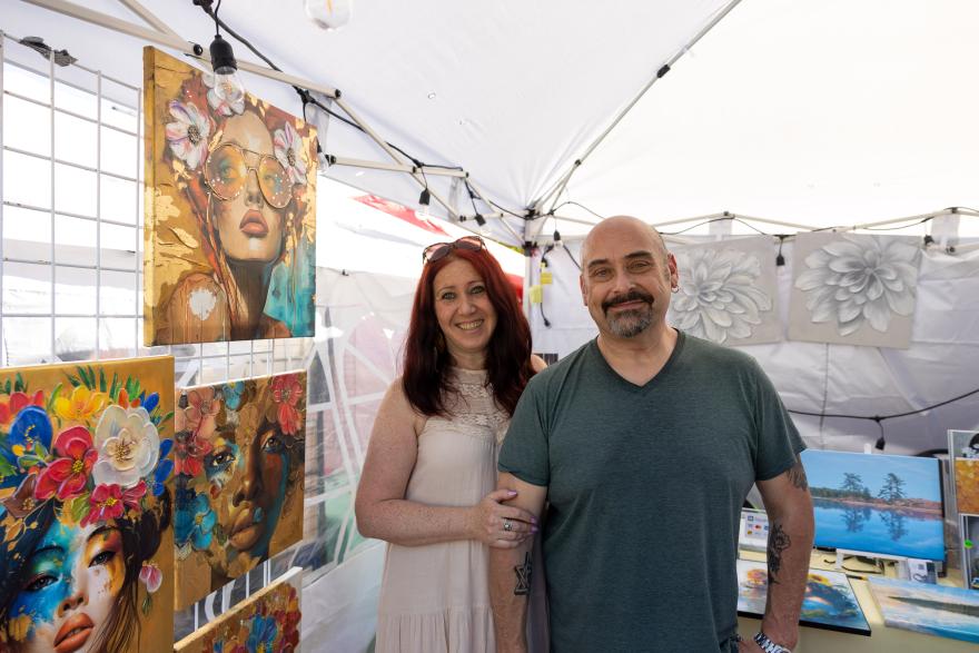 Man and woman smiling in art booth with colorful paintings displayed.