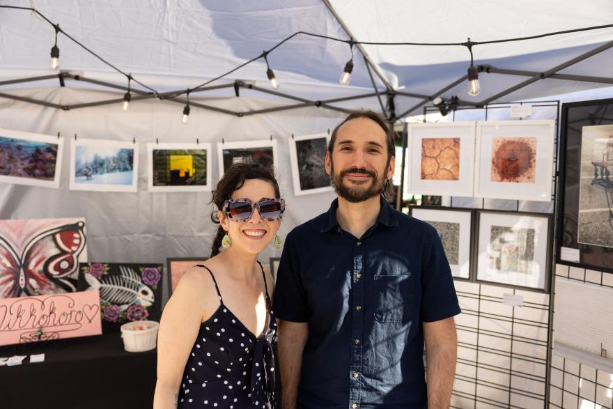 Couple smiling in an art booth with framed artworks behind them.