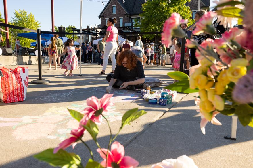 Woman drawing chalk art on pavement at a public event, surrounded by flowers.