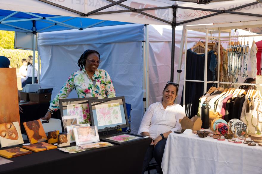 Market stall with two women, art displays, and clothes under a tent.