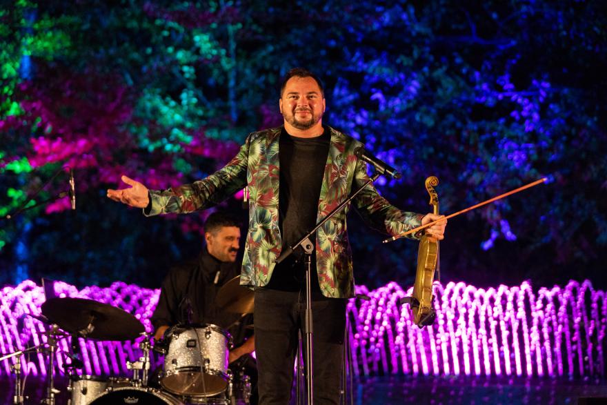 Musician with violin and microphone, colorful lights in background.