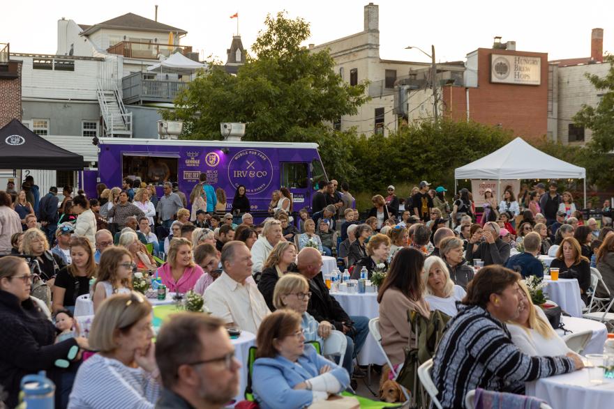 Crowd seated outdoors at an event with a purple food truck in the background.