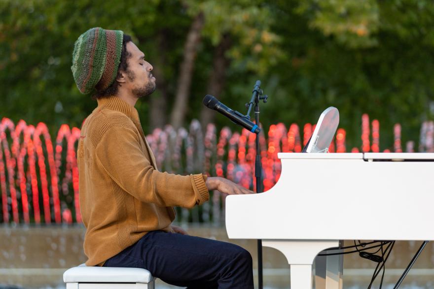 Man playing a white piano outdoors with colorful fountains in the background.