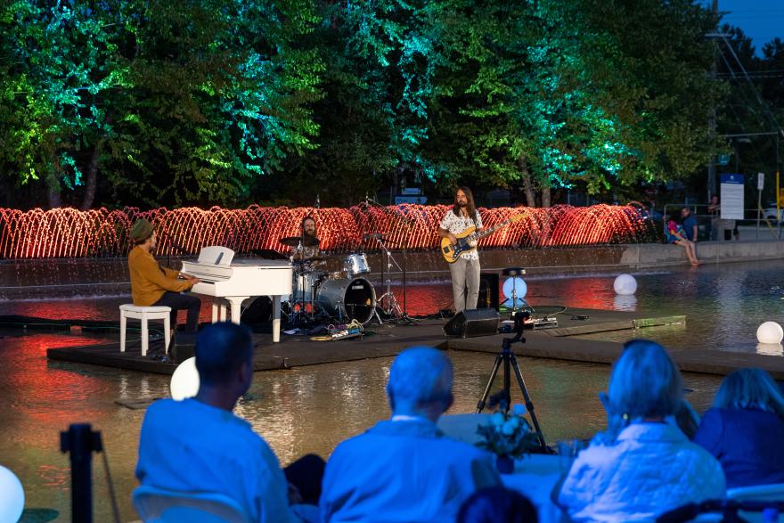 Outdoor concert with musicians on a stage over water, surrounded by colorful lights.