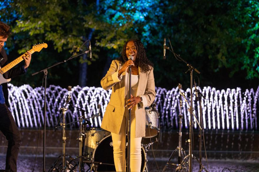Singer performing on stage, colorful lights and fountain in the background.