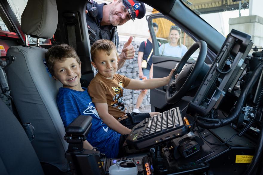 Two children sit inside a police car, smiling, with an officer standing nearby.