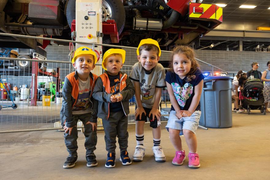 Four children wearing hard hats and safety vests stand in a garage setting, smiling.