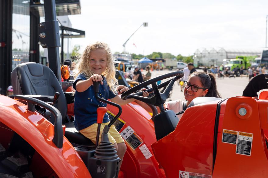 Child smiling while sitting on a red tractor, with an adult nearby at an outdoor event.