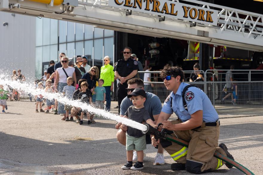 Firefighter helps child hold hose, spraying water. Crowd watches nearby.