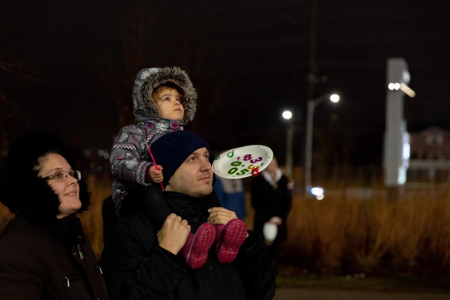Family watching lights at night; child on shoulders, bundled in warm clothes.