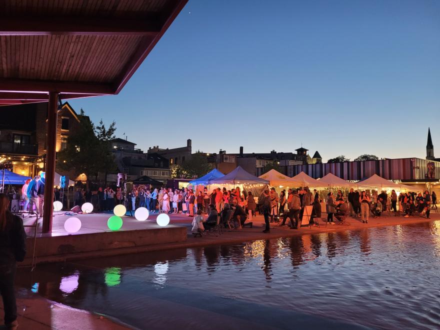 Outdoor evening market with tents and colorful lights by a pond, under a twilight sky.