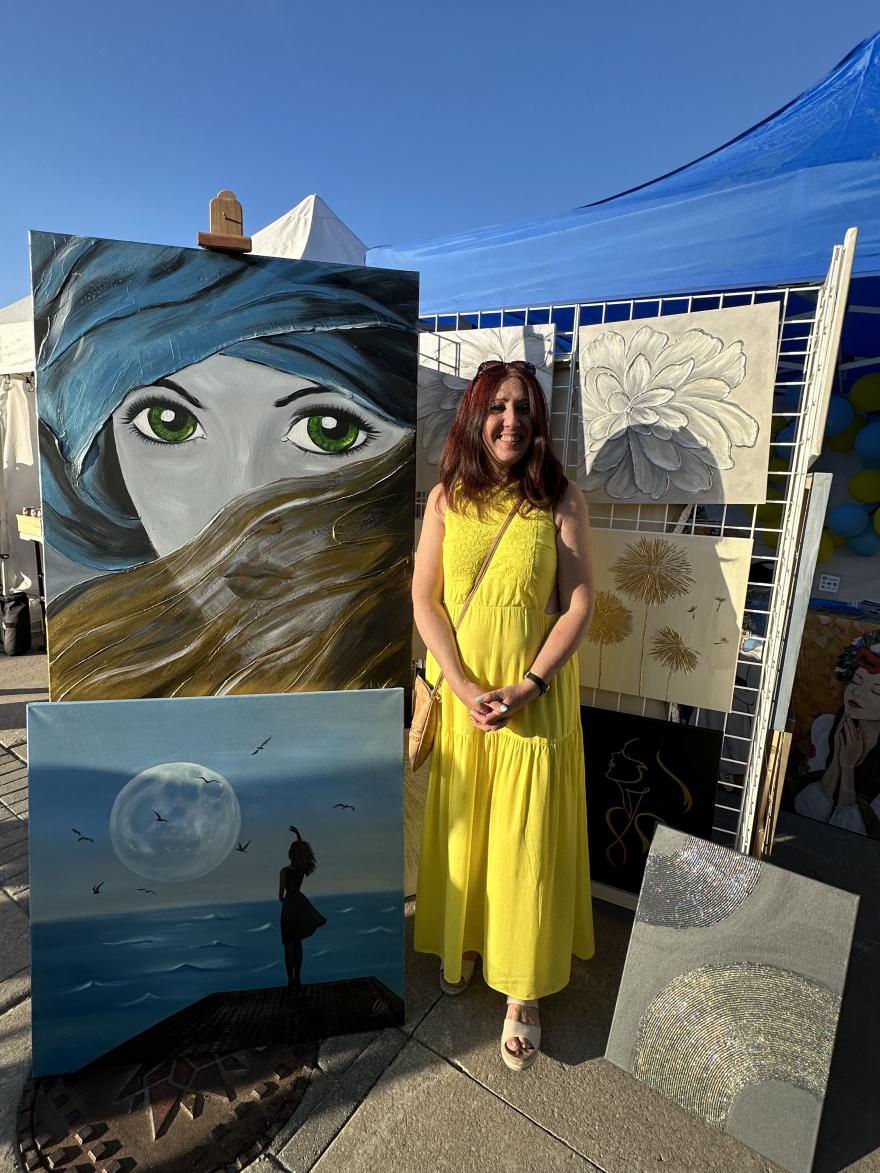 Woman in a yellow dress stands by her paintings at an outdoor art display.