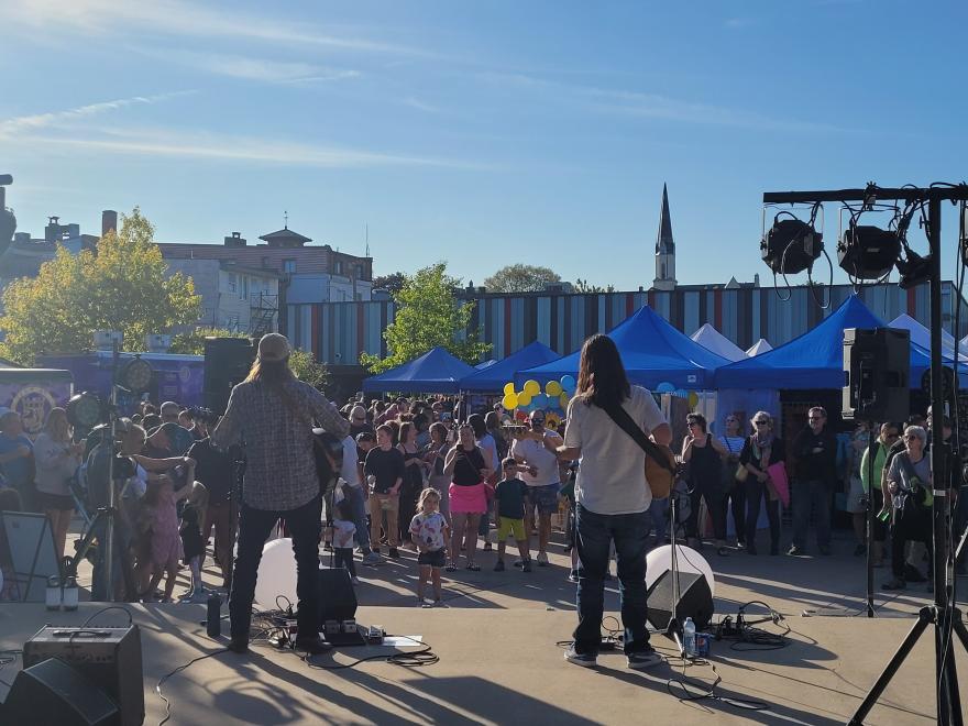 Musicians perform onstage outdoors to a crowd under blue tents.