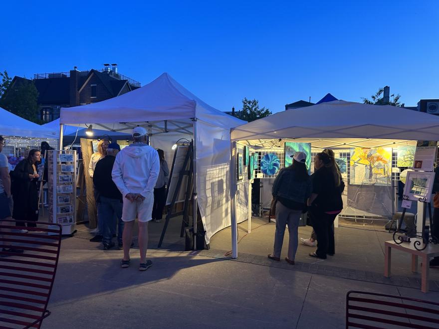 Outdoor art market at dusk with lit tents and visitors browsing.