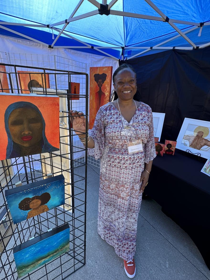 Artist smiles beside vibrant paintings at an art booth under a blue canopy.