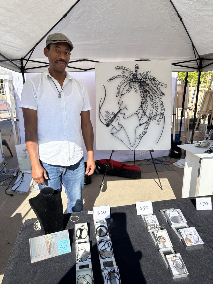 Man standing by art display booth under a tent at an outdoor market.