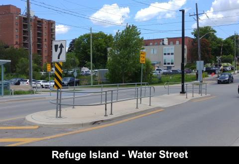 Refuge island on a city street, surrounded by buildings and greenery.