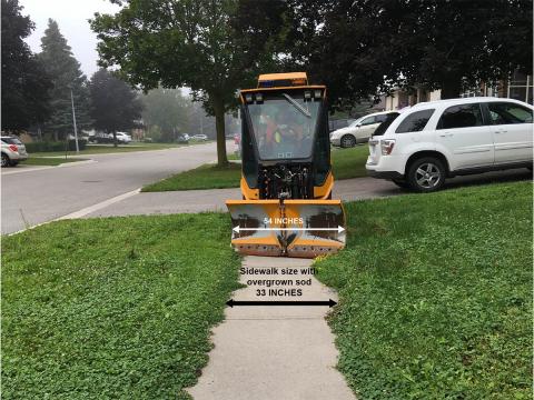 Small snowplow on a sidewalk near parked cars and trees.