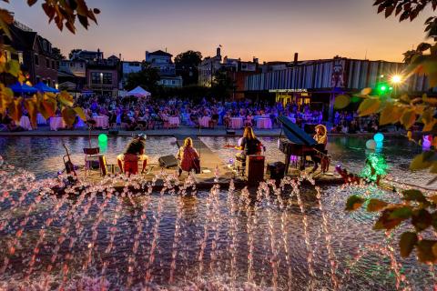 Open-air concert in a park at sunset, with vibrant lights and water fountains.