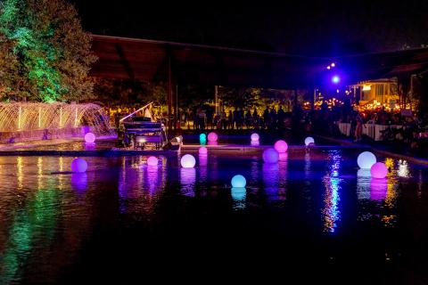 Glowing orbs float on a reflective pool at night, with a grand piano nearby.