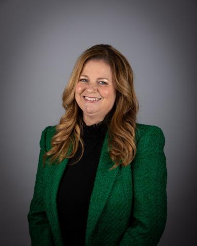 Kelly Broome smiling in a green jacket against a gray background.