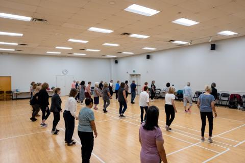 A group of adults participate in a line dance class in a large community hall